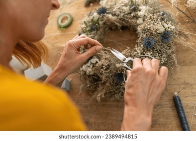 Close-up of a florist's hands using scissors to work on a dried flower wreath on a wooden table