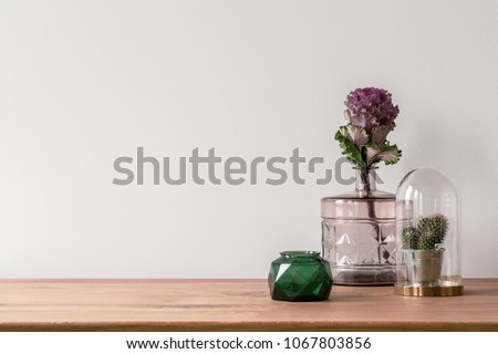 Close-up of a flower in a pink glass vase and a small cactus in a dome on the side of a wooden surface and an empty, white background
