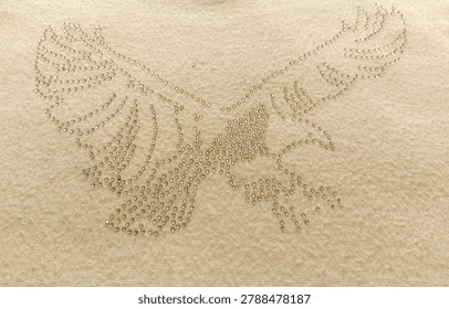 Close-up of a flying eagle shape made of shiny rhinestones on beige fabric, creating a luxury crystal embellishment pattern background for fashion design.