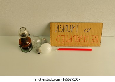 Closeup and focus on cardboard sign with handwriting DISRUPT or BE DISRUPTED placed on table among candle, oil lamp, incandescent lamp and LED lamp, disruptive technology concept