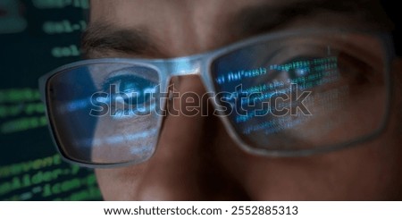A close-up of a focused programmer's face wearing eyeglasses which reflect lines of glowing green and blue programming code. Hacker, cybersecurity and AI concept