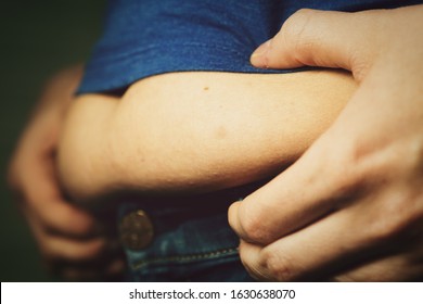 Close-up of a fold of fat sticking out from under a blue T-shirt of a woman in navy blue jeans, shallow depth of field, selective Focus. - Shutterstock ID 1630638070