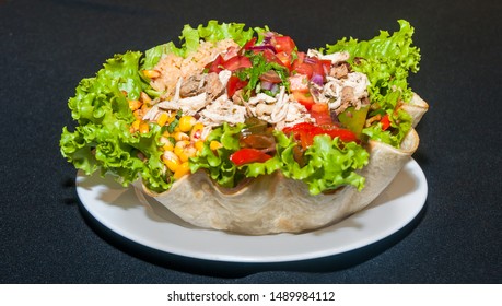 Closeup, Food Photography Picture Of A Burrito Bowl Filled With Chicken, Corn, Diced Tomatoes, Chilli Pepper, Lettuce And Rice, Tortilla Made Into A Bowl. White Plate On A Black Background