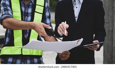 Close-up of a foreman and manager reviewing construction plans on-site, using a walkie-talkie and pen pointing at blueprint, Collaborative decision-making and modern communication in construction. - Powered by Shutterstock