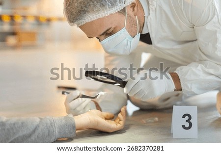Closeup of forensic expert examining corpse hand of dead body with magnifying glass collecting evidence. Criminologist in white safety suit inspecting crime scene. Murder investigation concept.