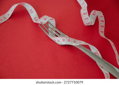 Closeup fork wrapped with long measuring tape on red background representing diet pressure, calorie restriction, weight loss routine and strict eating discipline in minimal symbolic composition