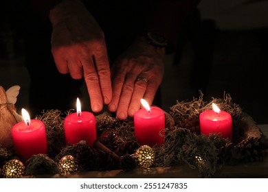 A close-up of four red advent candles with flames, surrounded by pinecones, moss, and small golden ornaments. A hand, wearing a wedding ring, is gently placed near the candles.  - Powered by Shutterstock