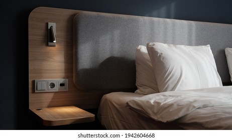 Close-up fragment of bedroom with empty bedside table, reading lamp and a USB socket in modern interior​ design home or hotel. Soft pillow and blanket, stylish comfortable furniture. Sun shadows.