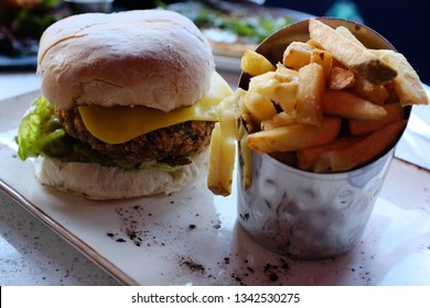 Closeup Of French Fries / Potato Chips In A Metal Pail Container And Blaa Bread Veggie Burger With Lettuce And Cheese On A Plate, Focused On The Burger