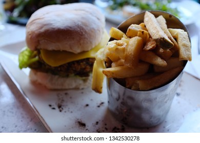 Closeup Of French Fries / Potato Chips In A Metal Pail Container And Blaa Bread Veggie Burger With Lettuce And Cheese On A Plate, Focused On The Fries