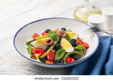 Close-up Of French Nicoise Salad With Anchovy, Green Beans, Tomatoes, Eggs, Black Olives, Capers On White Plate And White Wooden Background. Healthy Food, No Carb Diet Concept.