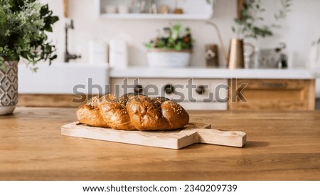closeup of fresh baked loaf bread or bun with sesame seeds on wooden cutting board and vase with flowers on wooden dining table on blurred kitchen background. homemade pastry concept