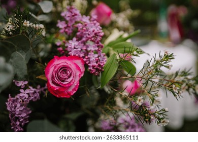 Close-up of a fresh pink rose with raindrops, surrounded by lilacs, greenery, and white blossoms in a vibrant floral arrangement.