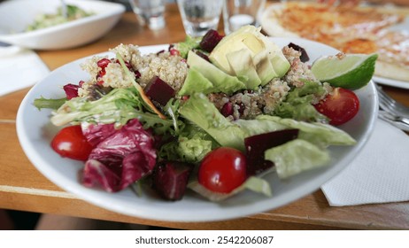 Close-up of a fresh and vibrant quinoa salad with mixed greens, avocado, cherry tomatoes, lime wedge, and beetroot on a dining table at a restaurant - Powered by Shutterstock