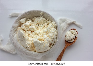 Close-up of freshly made white soft cottage cheese in bowl and wooden spoon on white table background. Homemade ricotta in cheesecloth. Top view. Fermented food with probiotics
