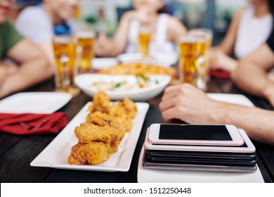 Close-up Of Fried Chicken On The Plate With Stack Of Mobile Phones Near By On The Wooden Table In Cafe With Young People In The Background