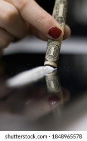 A close-up front image of a woman cocaine addict, holding a rolled dollar bill with her fingertips, to snort the cocaine powder strip that's on the mirror