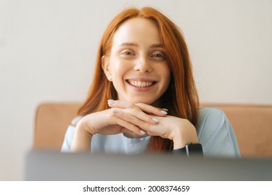 Close-up front view face of cheerful attractive young woman in casual clothing siting at desk with laptop, looking at camera, selective focus. Pretty redhead Caucasian lady remote working or studying.