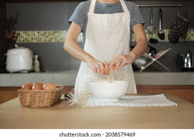 Close-up front view footage, a female cook in a white apron is cracking an egg into a cup to prepare a meal on a wooden table in the home's kitchen. Eating egg yolks is a healthy breakfast. - Powered by Shutterstock