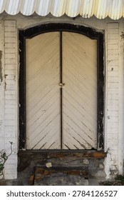 A close-up front view of an old, rustic wooden double door with a classic arch top design, set within a weathered white brick wall. The door features diagonal wood paneling and a vintage metal latch, evoking a sense of history, traditional architecture, and antique charm