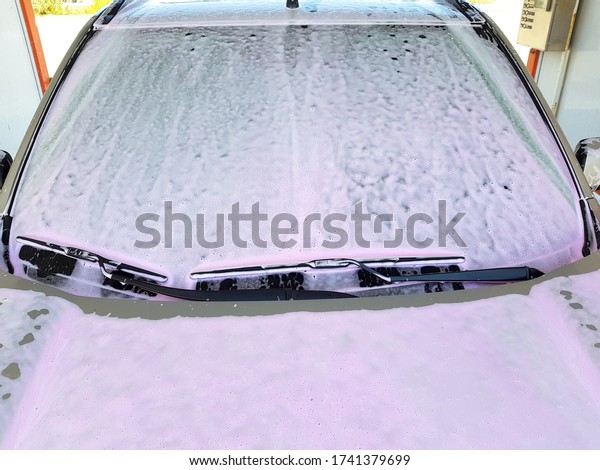 Close-up of the front windshield with wiper blades \
covered a pink washing active foam for cleanup a car at the\
self-service car wash.
