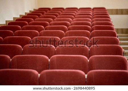 A close-up, front-on view of the empty maroon seats in a university lecture hall. The soft, upholstered chairs are arranged in neat rows, with wooden steps visible on the side.