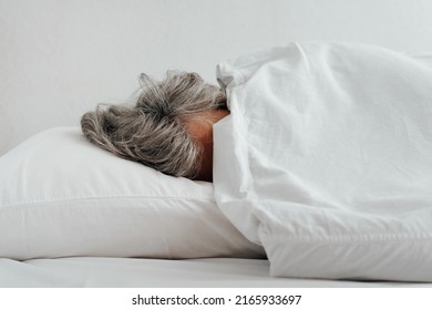Close-up of frozen caucasian woman with gray hair wrapped up in blanket lying on bed in bedroom. Low temperature, uncomfortable concept.
