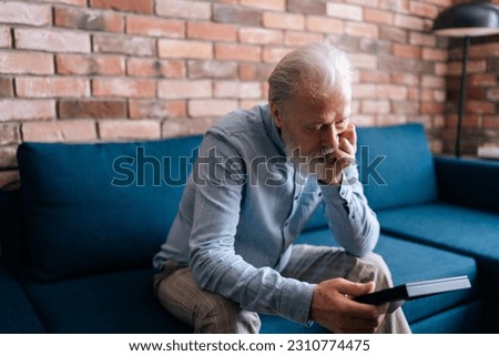 Closeup of frustrated senior male looking at frame of photo, stroking picture with sadness from missing someone, sitting on sofa at home. Concept of nostalgia, grief, longing and loneliness in old age