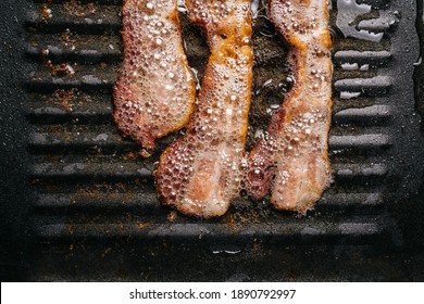 Close-up frying sizzling bacon strips in a pan with hot greasing bacon oil for making crispy bacon. Keto snack meal.