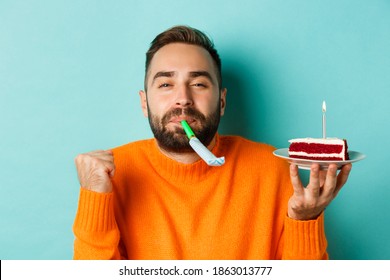 Close-up of funny adult man celebrating his birthday, holding bday cake with candle, blowing party wistle and rejoicing, standing over light blue background