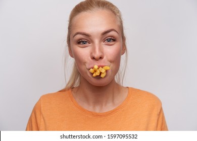 Close-up of funny attractive young blonde female with casual hairstyle looking joyfully at camera and having mouth full of french fries, fooling with food while posing over white background