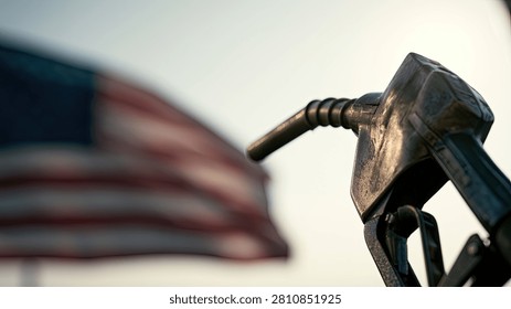 A close-up of a gas pump nozzle in front of a blurred American flag, representing the cost and availability of fuel in the United States.