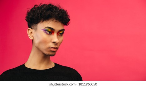 Close-up Of A Gender Fluid Man With Rainbow Eye Makeup Looking Away On Red Background. Transgender Male With Pride Flag Eye Shadow.