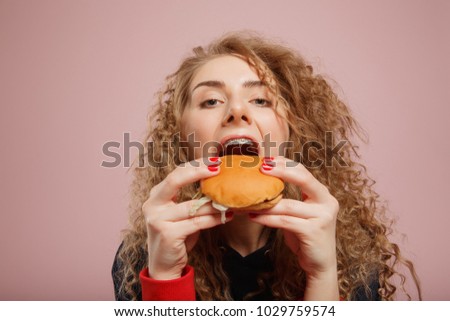 Close-up of girl with braces on her teeth and curly hair biting juicy burger, pink background. Concept fast food, hunger, teenager