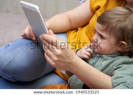 Close-up of a girl in her mother's arms watching a tablet. Mom is working at home while taking care of her daughter. Concept of technology and conciliation.