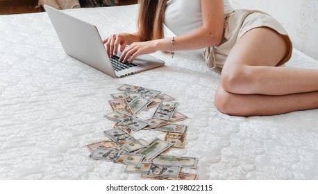 A Close-up Of A Girl Lying On A Mattress Behind A Laptop Against The Background Of Scattered Dollar Money.Freelancer Earns At Home,profitable Business,remote Work.A Rich Woman Enjoys Dollar Bills.Cash