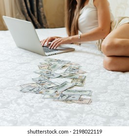 A Close-up Of A Girl Lying On A Mattress Behind A Laptop Against The Background Of Scattered Dollar Money. A Freelancer Earns At Home, A Profitable Business. A Rich Woman Enjoys Dollar Bills. Cash