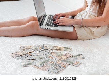 A Close-up Of A Girl Lying On A Mattress Behind A Laptop Against The Background Of Scattered Dollar Money.Freelancer Earns At Home,profitable Business,remote Work.A Rich Woman Enjoys Dollar Bills.Cash