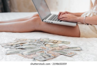 A Close-up Of A Girl Lying On A Mattress Behind A Laptop Against The Background Of Scattered Dollar Money.Freelancer Earns At Home,profitable Business,remote Work.A Rich Woman Enjoys Dollar Bills.Cash