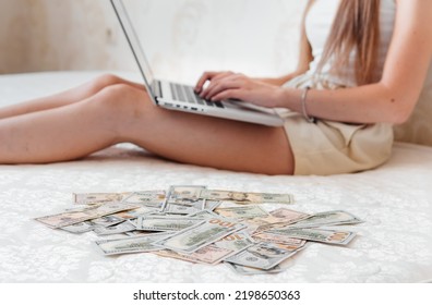 A Close-up Of A Girl Lying On A Mattress Behind A Laptop Against The Background Of Scattered Dollar Money.Freelancer Earns At Home,profitable Business,remote Work.A Rich Woman Enjoys Dollar Bills.Cash