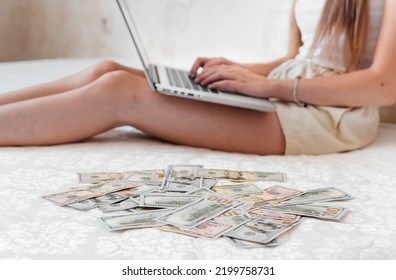 A Close-up Of A Girl Lying On A Mattress Behind A Laptop Against The Background Of Scattered Dollar Money.Freelancer Earns At Home,profitable Business,remote Work.A Rich Woman Enjoys Dollar Bills.Cash