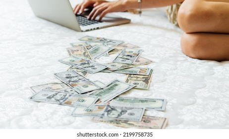 A Close-up Of A Girl Lying On A Mattress Behind A Laptop Against The Background Of Scattered Dollar Money. A Freelancer Earns At Home, A Profitable Business. A Rich Woman Enjoys Dollar Bills. Cash