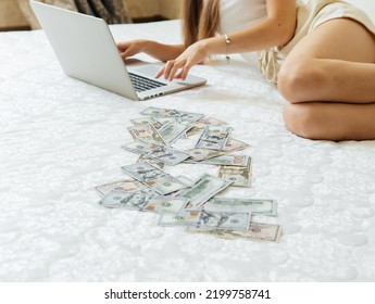 A Close-up Of A Girl Lying On A Mattress Behind A Laptop Against The Background Of Scattered Dollar Money.Freelancer Earns At Home,profitable Business,remote Work.A Rich Woman Enjoys Dollar Bills.Cash