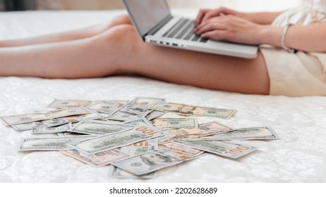 A Close-up Of A Girl Lying On A Mattress Behind A Laptop Against The Background Of Scattered Dollar Money.Freelancer Earns At Home,profitable Business,remote Work.A Rich Woman Enjoys Dollar Bills.Cash