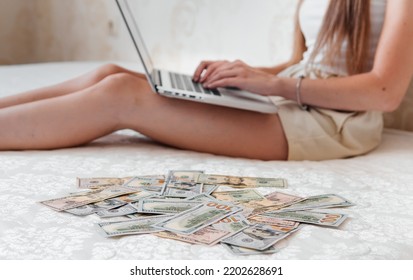 A Close-up Of A Girl Lying On A Mattress Behind A Laptop Against The Background Of Scattered Dollar Money.Freelancer Earns At Home,profitable Business,remote Work.A Rich Woman Enjoys Dollar Bills.Cash