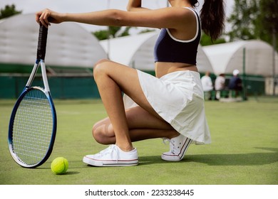 Close-up of girl on tennis court. Woman knelt down on one knee, holding tennis racket in hand. She is wearing dark gray top, white skirt and white sneakers. Tennis ball is lying on the side. - Powered by Shutterstock