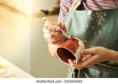 Close-up of girl painting clay mug with glaze. Woman coloring pottery in workshop with a paintbrush. Painter in green apron glazing clay pot.  - Powered by Shutterstock