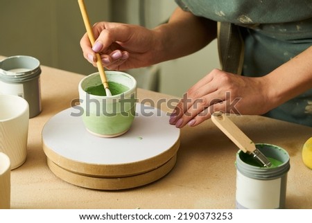 Close-up of girl painting clay mug with glaze. Woman coloring pottery in workshop with a paintbrush. Painter in green apron glazing clay pot. 