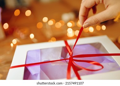 Close-up Of Girl's Hand Pulling Ribbon From Surprise Box. Bokeh Lights In Background. Unpacking Gift On Wooden Table. Festive Concept Of Christmas Or Birthday.