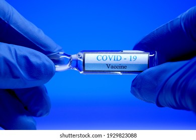 Close-up glass ampoule with covid-19 vaccine in the hands of a medical worker in blue sterile gloves on a blue background. Coronavirus vaccination concept